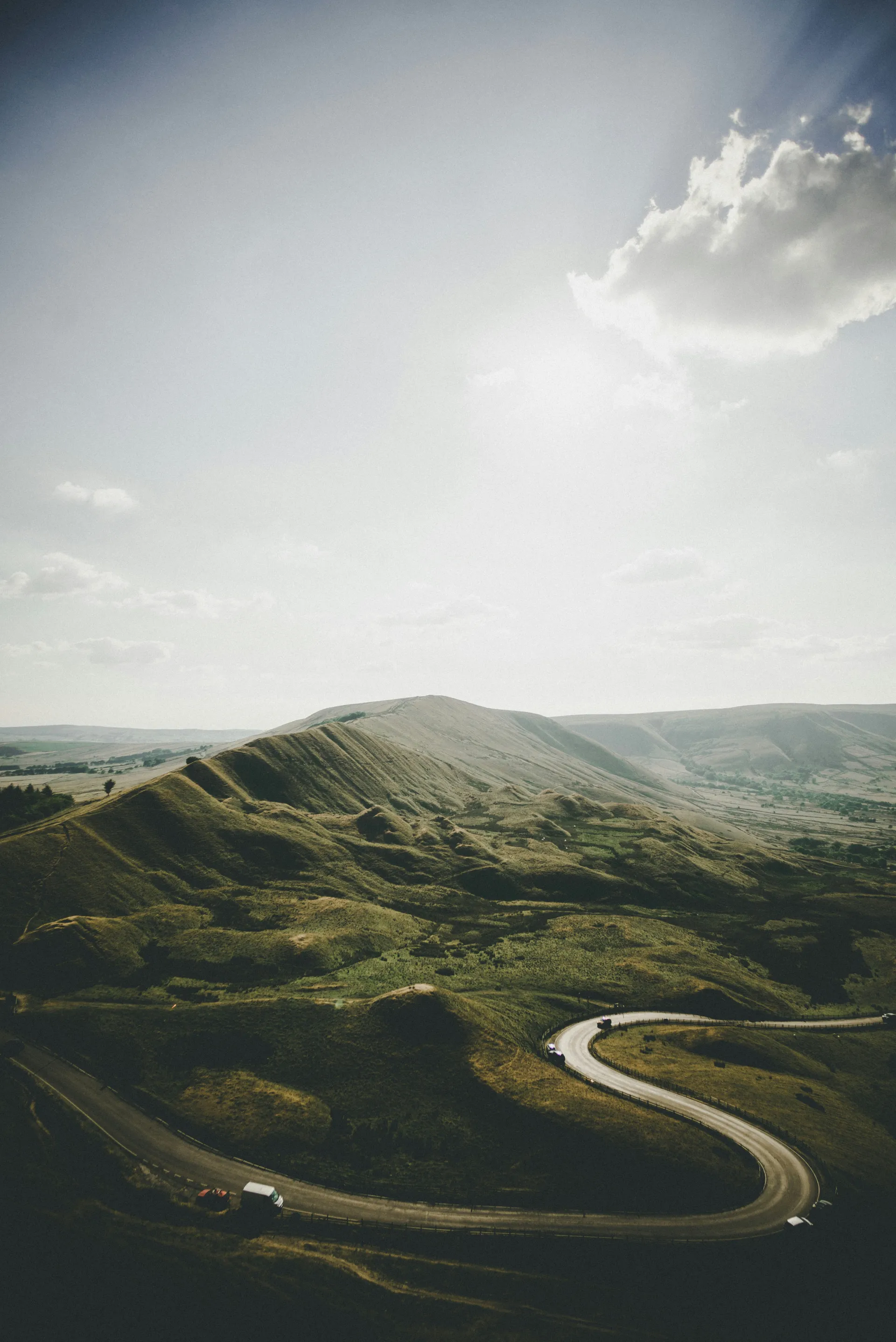 The winding road below Mam Tor in the Peak District, green hills under a wide sky