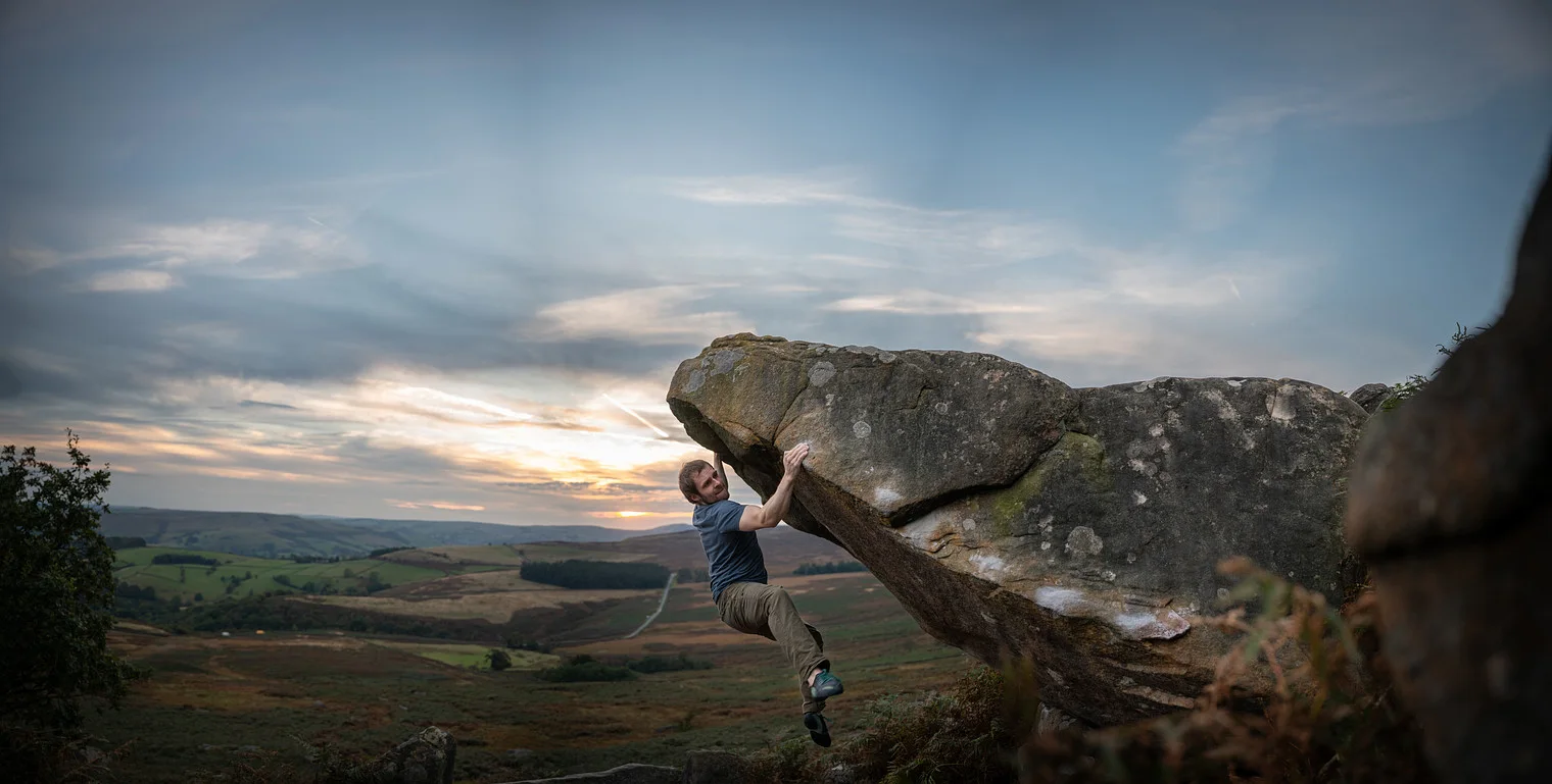 A boulderer topping out on a Stanage Edge gritstone block at sunset, the Peak District valley beyond